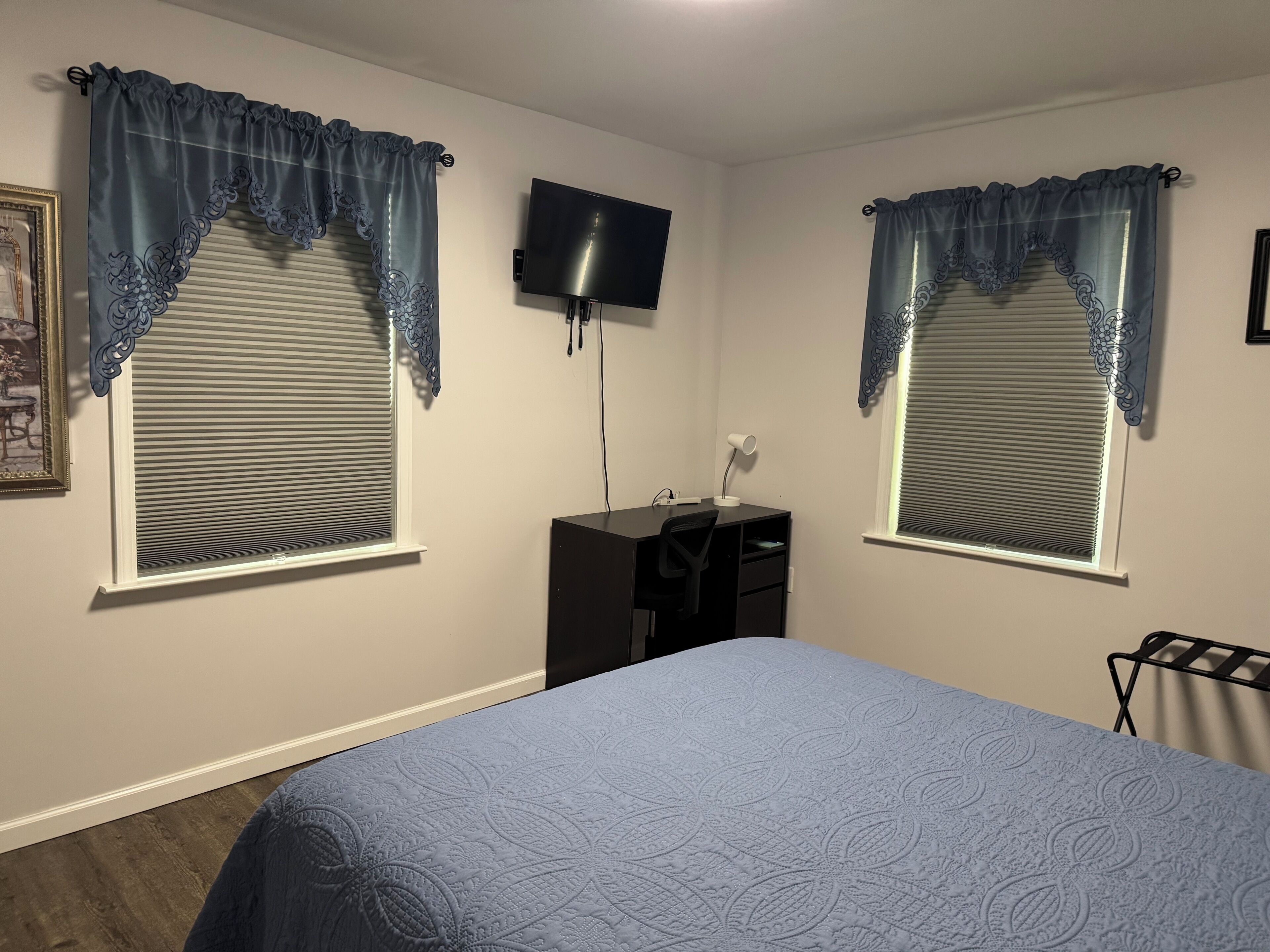 A bedroom with two windows, a bed, a desk with a chair, and a flat-screen TV mounted on the wall.