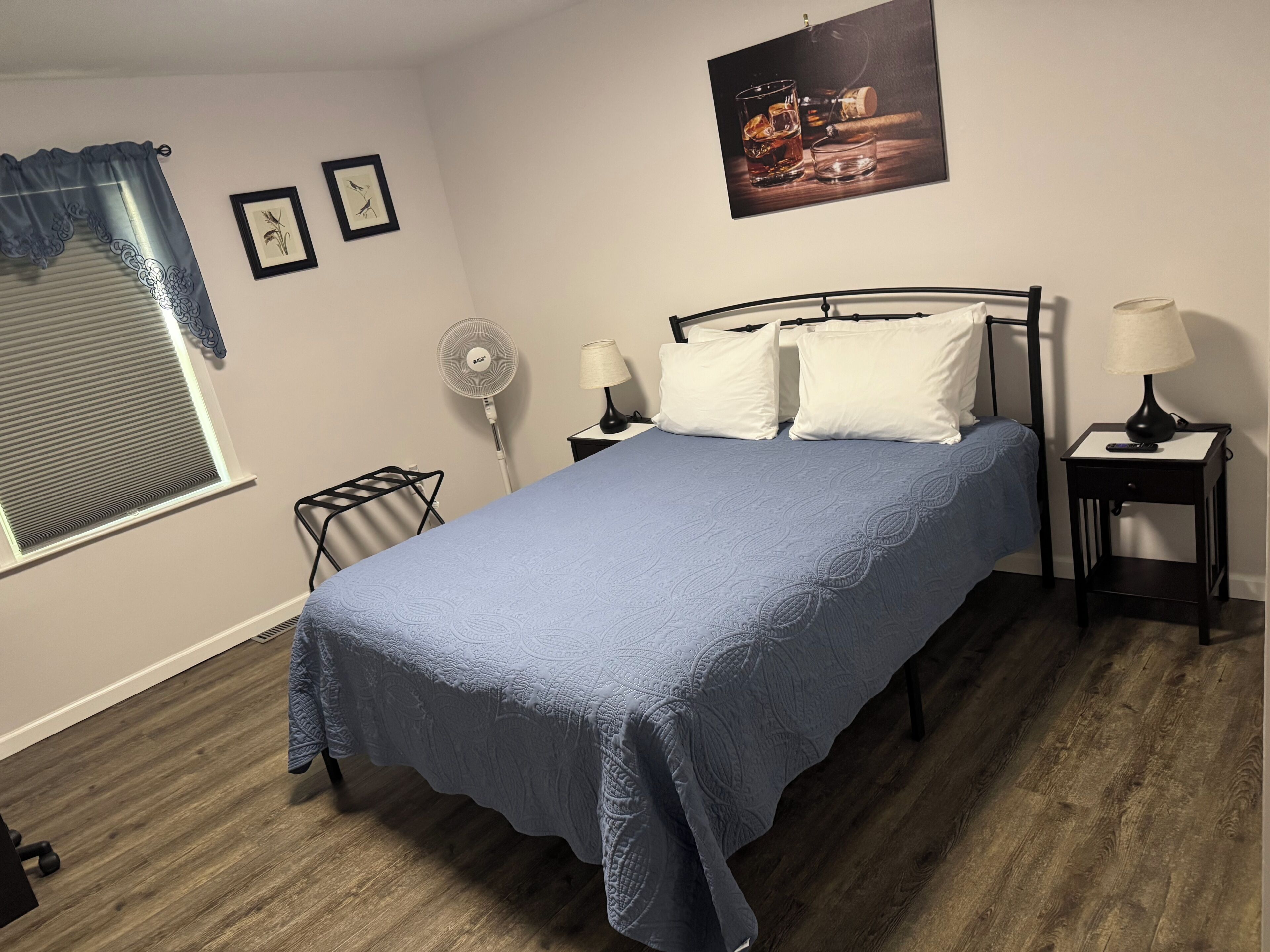 A bedroom with a bed, two bedside tables, a standing fan, a window with blinds, and framed pictures on the wall.