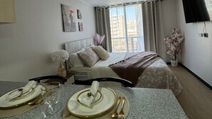 Une chambre d’hôtel avec un lit, une télévision, une table à manger dressée avec des assiettes et des couverts, et une fenêtre avec des rideaux.