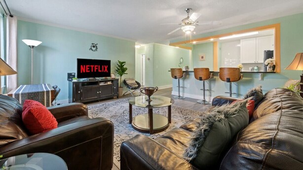 Ein Wohnzimmer mit Ledersofa, einem Glastisch, einem Fernseher, der 'NETFLIX' zeigt, und einer Küchenzeile im Hintergrund.