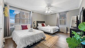 Ein Hotelzimmer mit zwei Betten, einem Deckenventilator, großen Fenstern mit Jalousien, einem Holzboden und einer Zimmerpflanze.