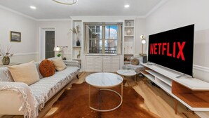 A modern living room with a large TV displaying "NETFLIX," a sofa, a coffee table, and a view of the city through a window.