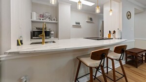 A modern kitchen with a white countertop, bar stools, pendant lights, and a microwave.