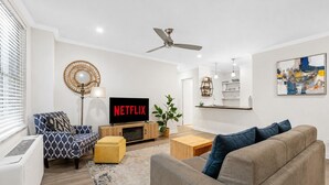 A modern living room with a flat-screen TV, a ceiling fan, a dining area, and a kitchen in the background.