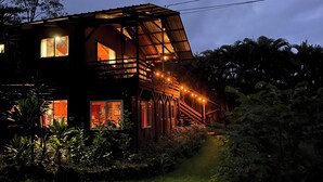 Uma casa de madeira de dois andares com varanda e luzes de Natal, cercada por vegetação exuberante.