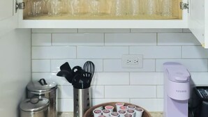 Una cocina con un mueble que contiene tazas blancas y copas de vino, una cafetera y una bandeja con cápsulas de café.