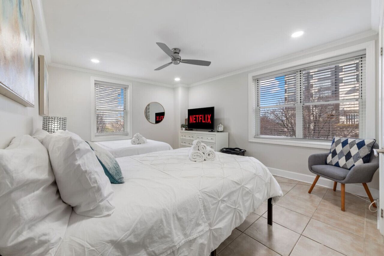 A bedroom with two beds, a ceiling fan, a TV displaying 'NETFLIX', a window with blinds, and a chair with a patterned cushion.
