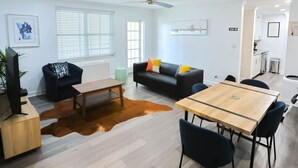 Un salon moderne avec un canapé, une table basse et une table à manger. On peut voir un téléviseur, une plante et un coin cuisine.