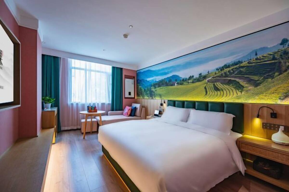 A hotel room with a large bed, a small table, a chair, and a painting of a landscape on the wall.