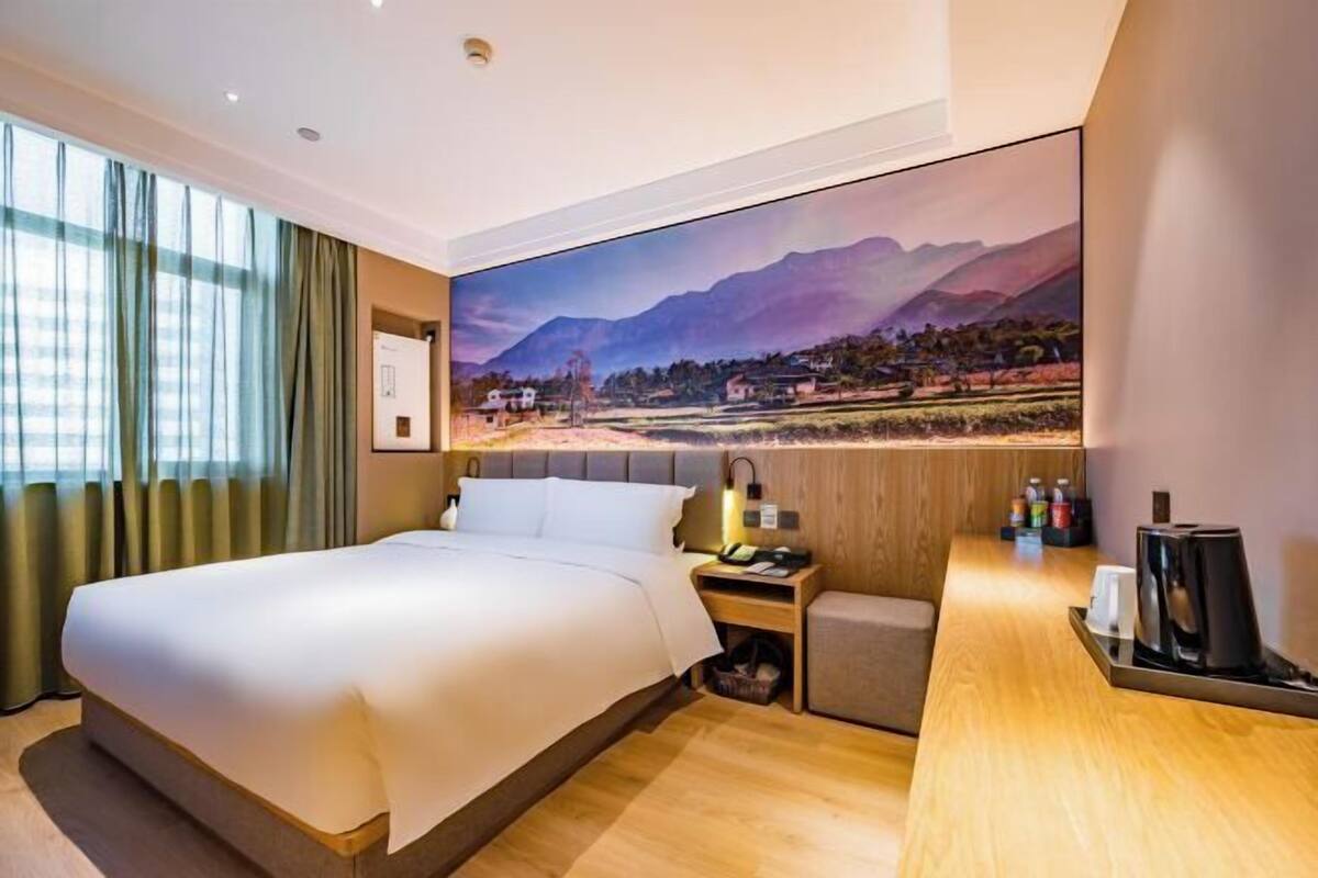 A hotel room with a large bed, a desk with a lamp, a small seating area, and a scenic mural on the wall.