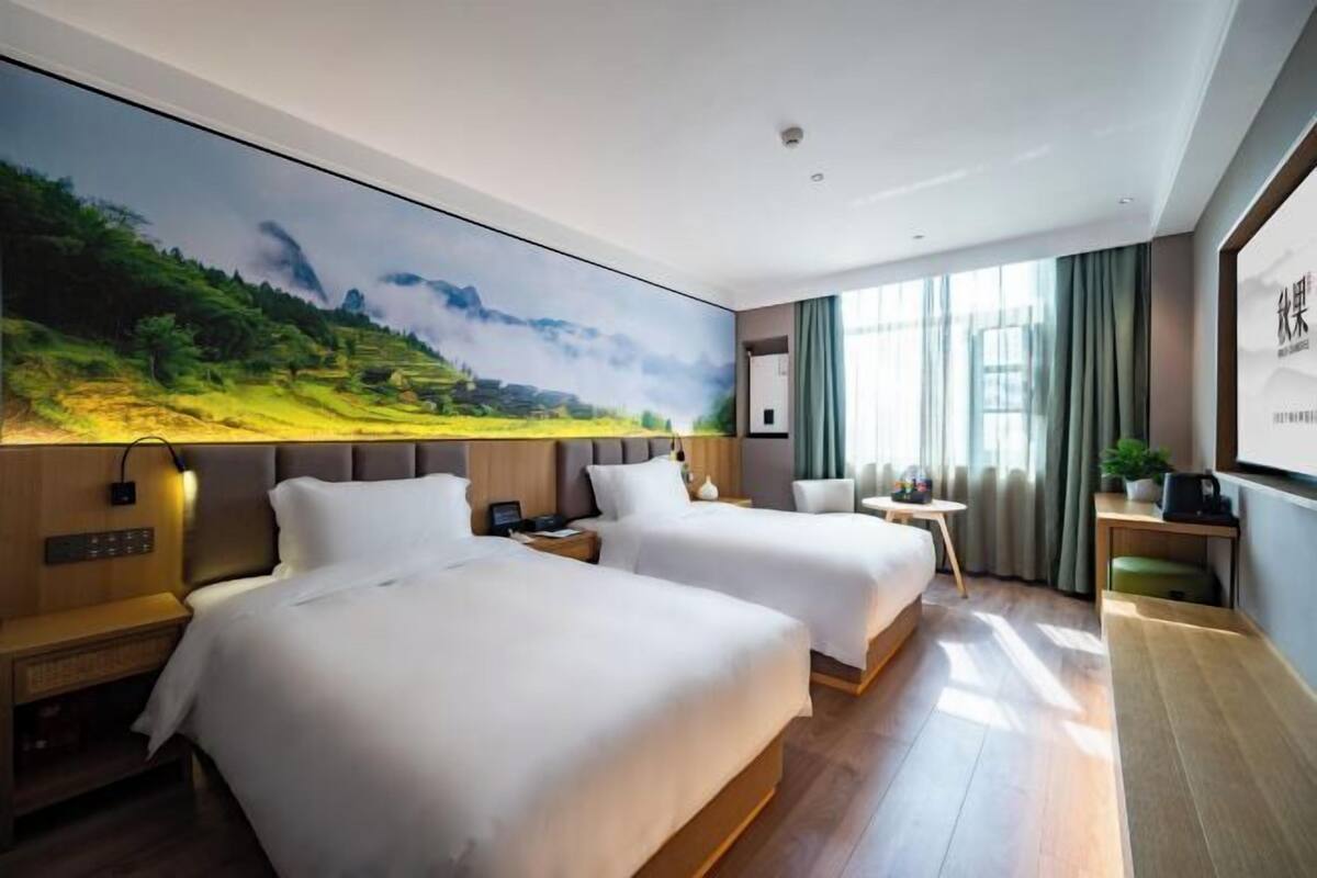 A hotel room with two beds, a large landscape painting on the wall, a desk with a chair, and a window with curtains.