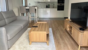 Una sala de estar moderna con un sofá gris, una mesa de centro de madera y un mueble para TV, que da paso a una cocina con armarios y electrodomésticos blancos.