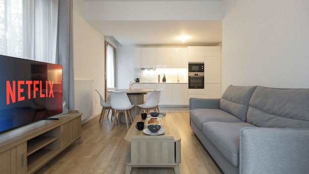 Una sala moderna con un televisor de pantalla plana, un sofá gris, una mesa de centro de madera y un comedor con sillas blancas y una pequeña mesa.