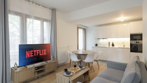 Una sala de estar moderna con un televisor de pantalla plana, un sofá gris, una mesa de centro de madera y un comedor con una mesa redonda y sillas blancas.