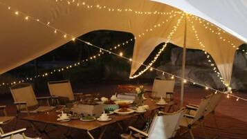Een buiten eethoek met een gedekte tafel, omringd door stoelen, onder een tent met sfeerverlichting.