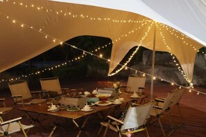 Un área de comedor al aire libre con una mesa lista para una comida, rodeada de sillas, bajo una carpa con guirnaldas de luces.