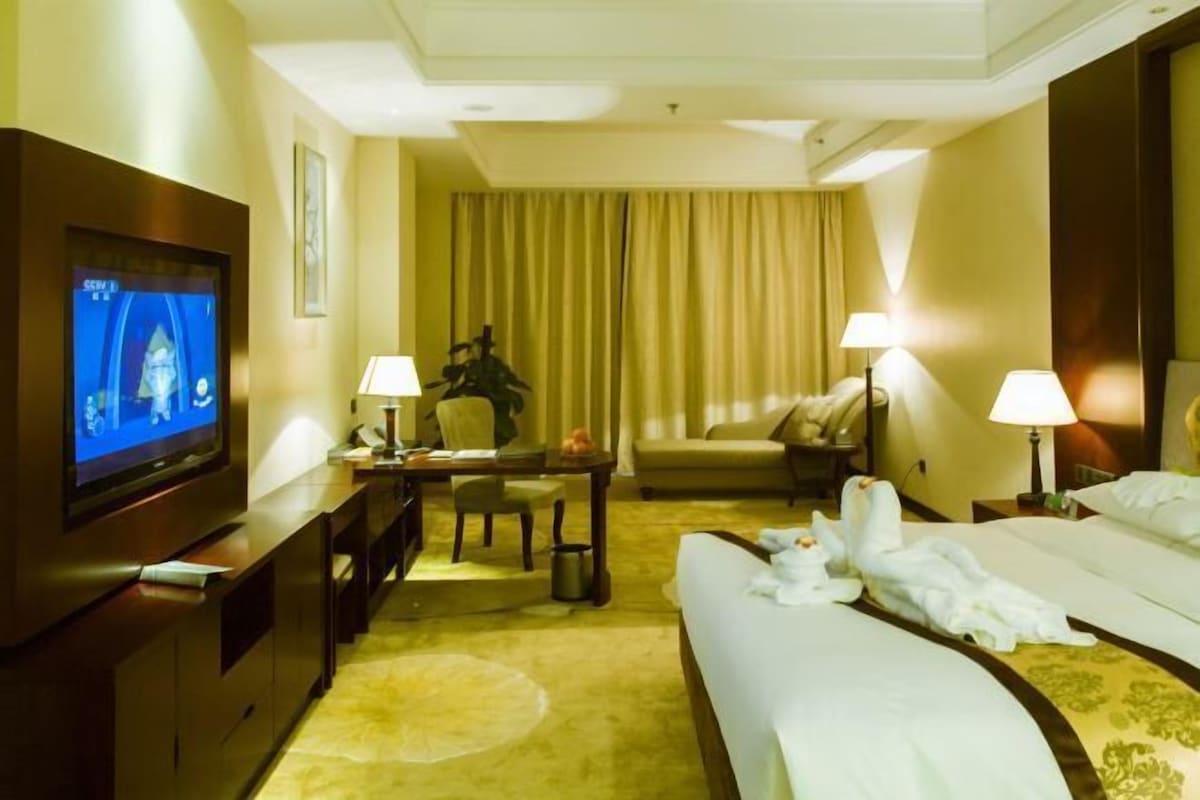 A hotel room with a large flat-screen TV, a desk with a lamp, a sofa, and two beds with towels.