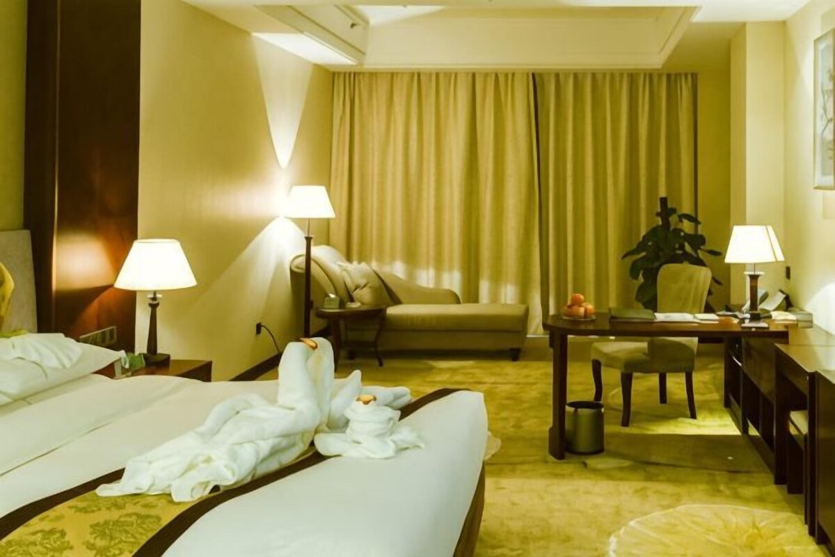 A hotel room with a bed, two bedside tables with lamps, a desk with a chair, a small table with a plant, and a large window with curtains.