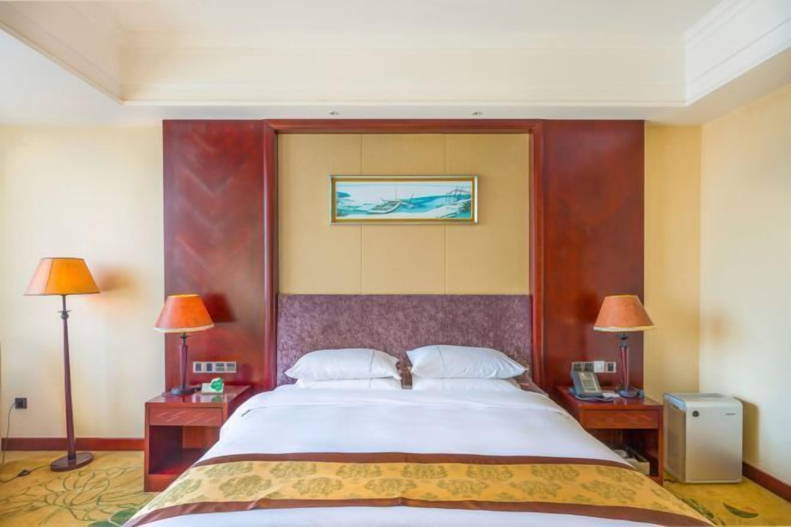 A hotel room with a bed, two bedside tables, a nightstand, a lamp, a small heater, and a painting on the wall.