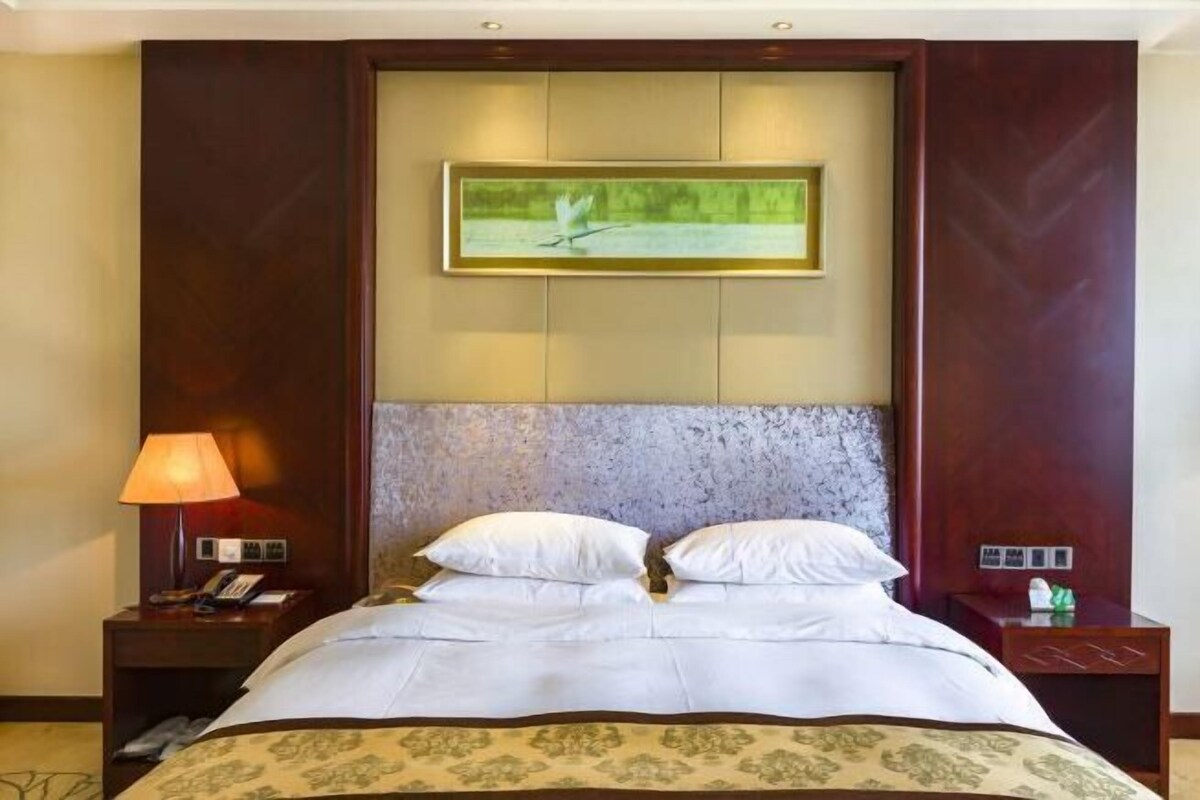 A hotel room with a large bed, a nightstand with a lamp, a framed picture on the wall, and a small table with a phone and a bottle.