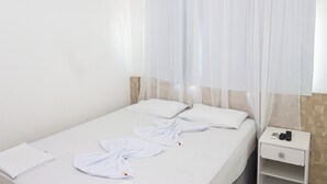 Habitación de hotel con cama, cortinas blancas, mesita de noche y un control remoto, además de una libreta.