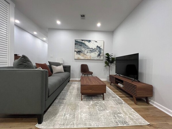 A modern living room with a grey sofa, a wooden coffee table, a TV stand, and a large abstract painting on the wall.