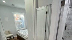 A bedroom with a wooden floor, a bed with a white and gray striped bedspread, a white door, and a window with blinds.