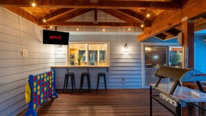A cozy outdoor area with a wooden deck, a bar with stools, a grill, and a TV screen displaying 'NETFLIX'. - Pool*Sauna*HotTub*Theater*Bar*Pickleball*Volleyball (Oakhurst)