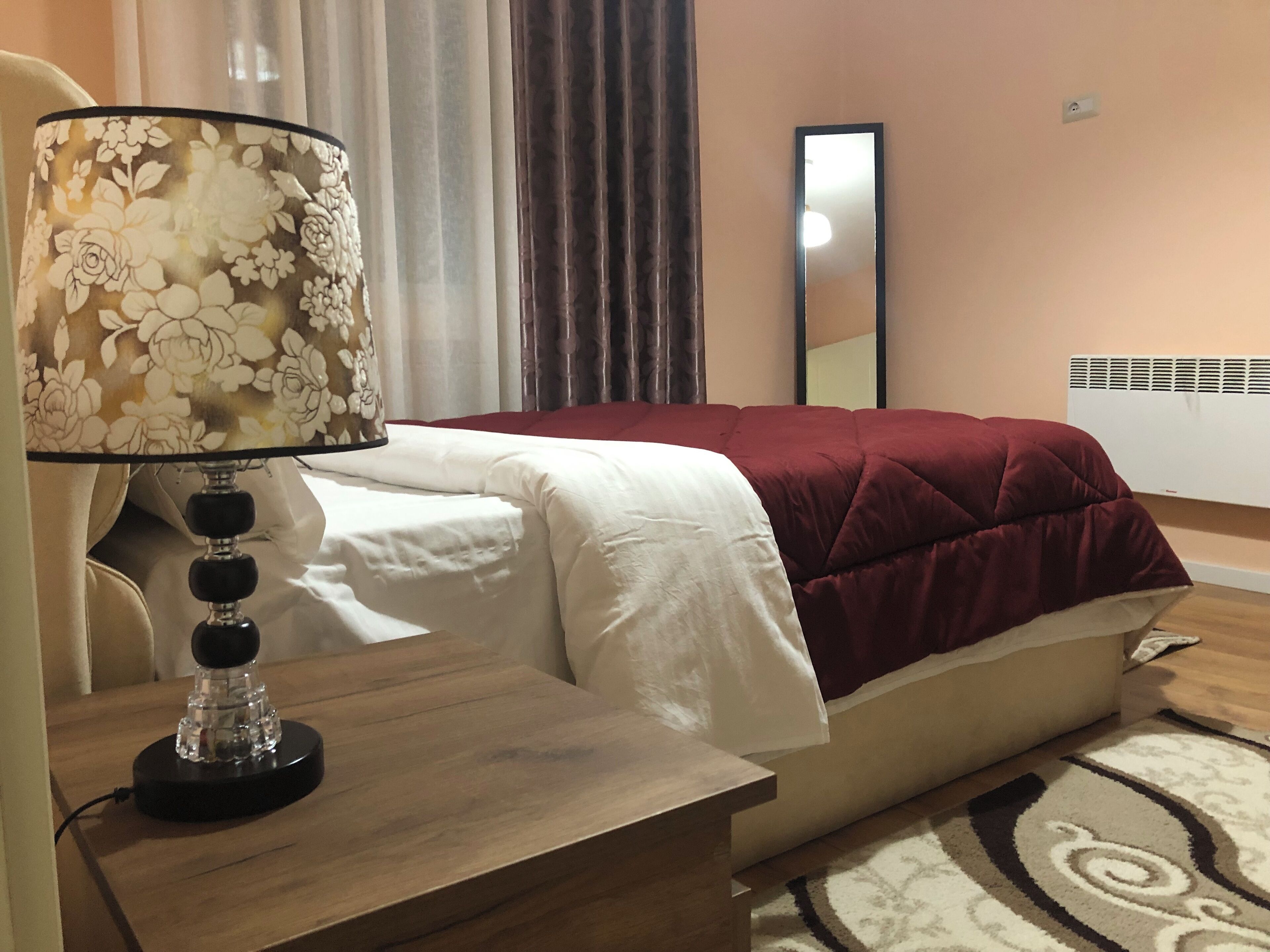 Un dormitorio ordenado con cama, mesita de noche con lámpara, espejo y radiador.