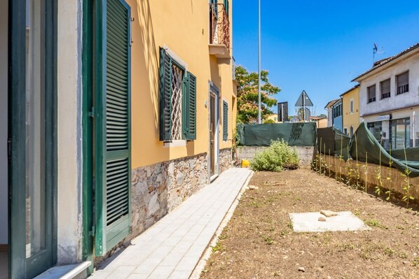 Une ruelle étroite avec un chemin pavé, un petit espace aménagé en jardin et un bâtiment aux volets verts et à la base en pierre.