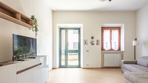 Un soggiorno con una televisione a schermo piatto, un divano, uno scaffale in legno con una pianta, una porta che dà sull'esterno e una finestra con tende.
