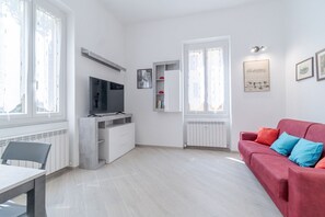 Uma sala de estar moderna com um sofá vermelho, uma televisão sobre um móvel, uma mesa de jantar e uma janela com cortinas.