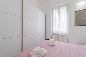 Quarto com colcha rosa, toalhas brancas, radiador e janela com cortinas.