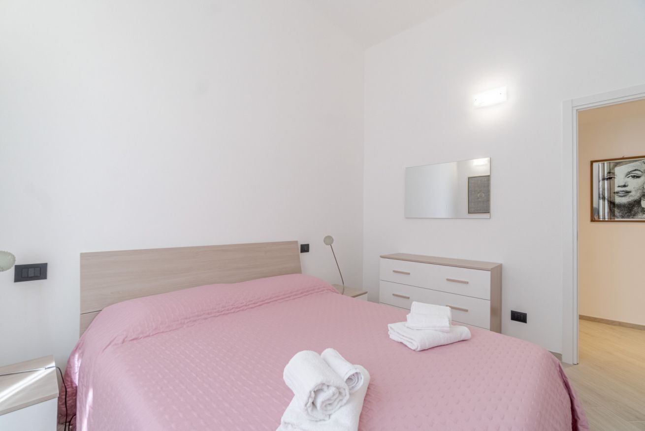 A bedroom with a bed, a bedside table, a dresser, a mirror, and a door leading to another room.