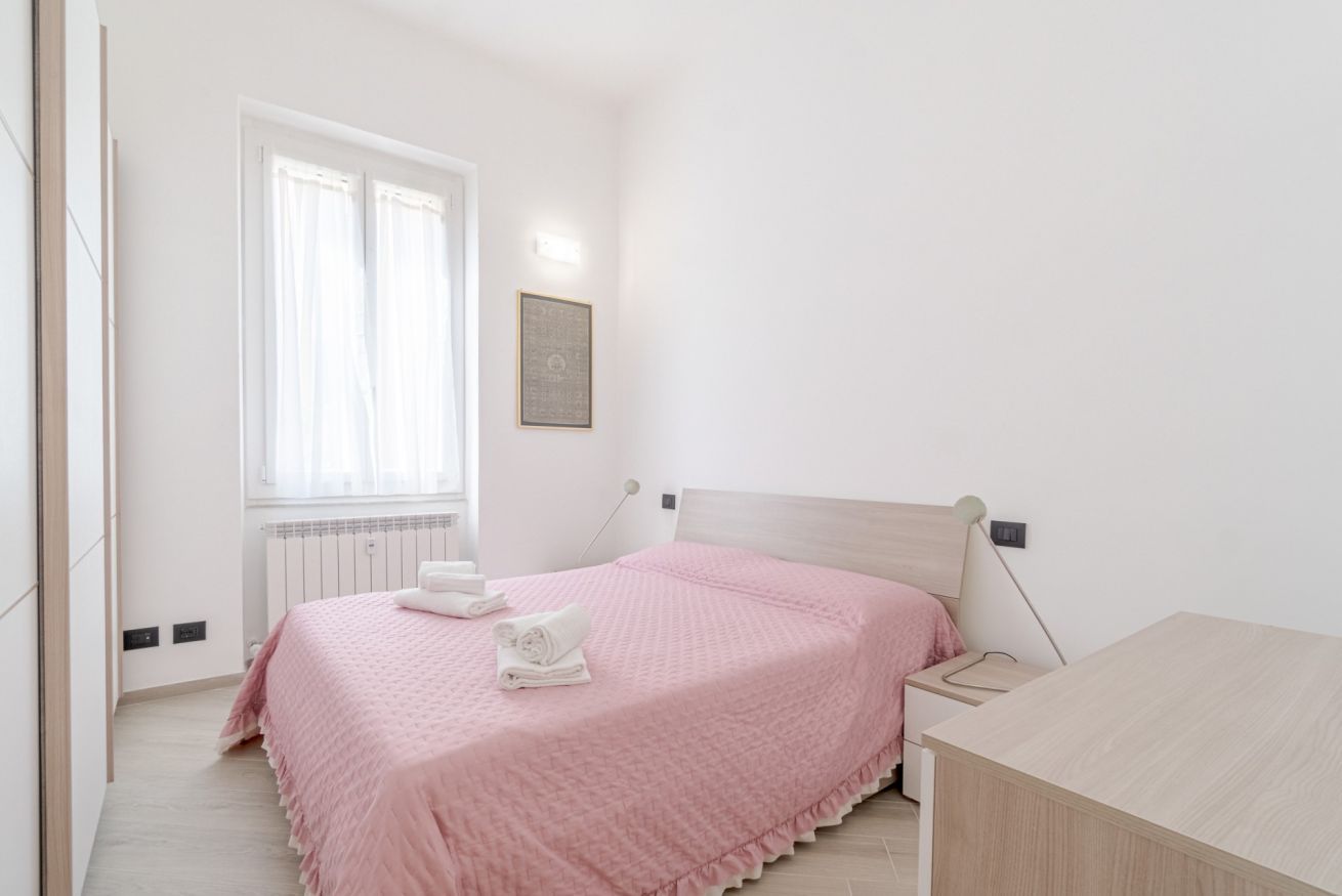 A bedroom with a bed, a bedside table, a window with curtains, a radiator, and a wall-mounted light.