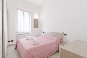 Et soveværelse med seng, sengebord, et vindue med gardiner, en radiator og en vægmonteret lampe.