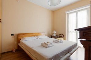 Une chambre d’hôtel avec un lit en bois, une table de chevet avec une lampe, une chaise et une vue sur l’extérieur par une fenêtre.