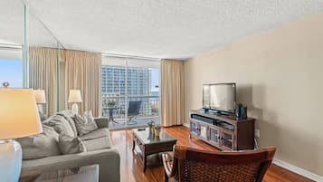A hotel room with a sofa, a television, a coffee table, and a view of a cityscape through large windows.