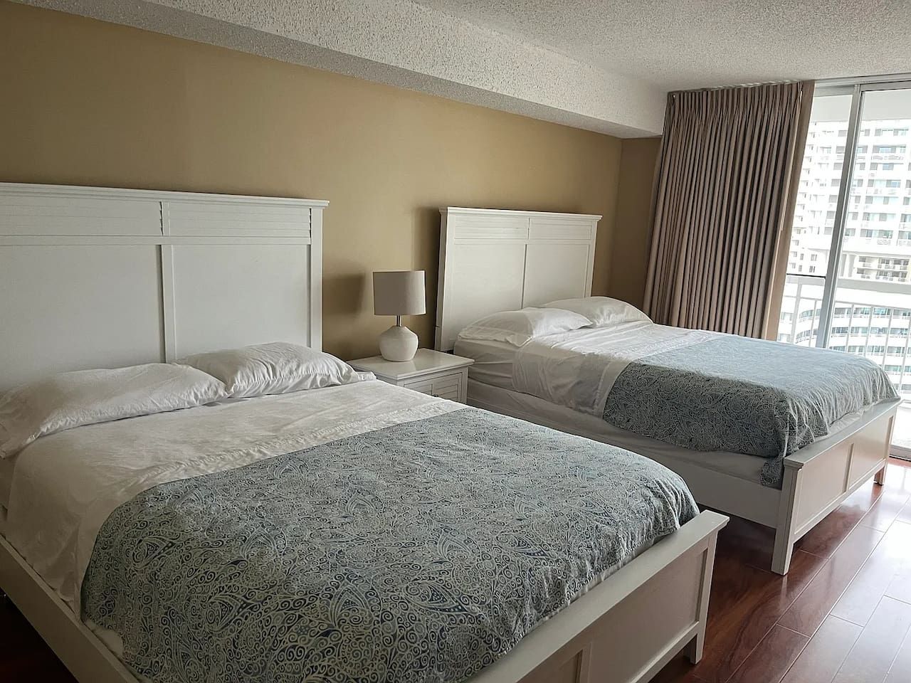 A hotel room with two beds, a nightstand with a lamp, and a large window with curtains.