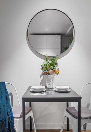 A dining area with a round mirror above a table, two chairs, a vase with flowers, and a blue jacket hanging on a chair.