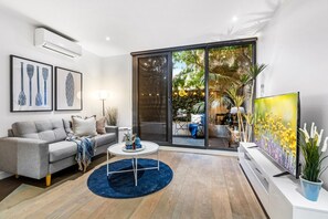 A modern living room with a grey sofa, a coffee table, and a TV. There is a large sliding glass door leading to an outdoor area with plants and a table.