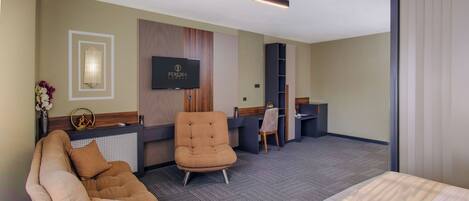 A hotel room with a sofa, armchair, and a bed. There is a TV mounted on the wall and a desk with a chair.