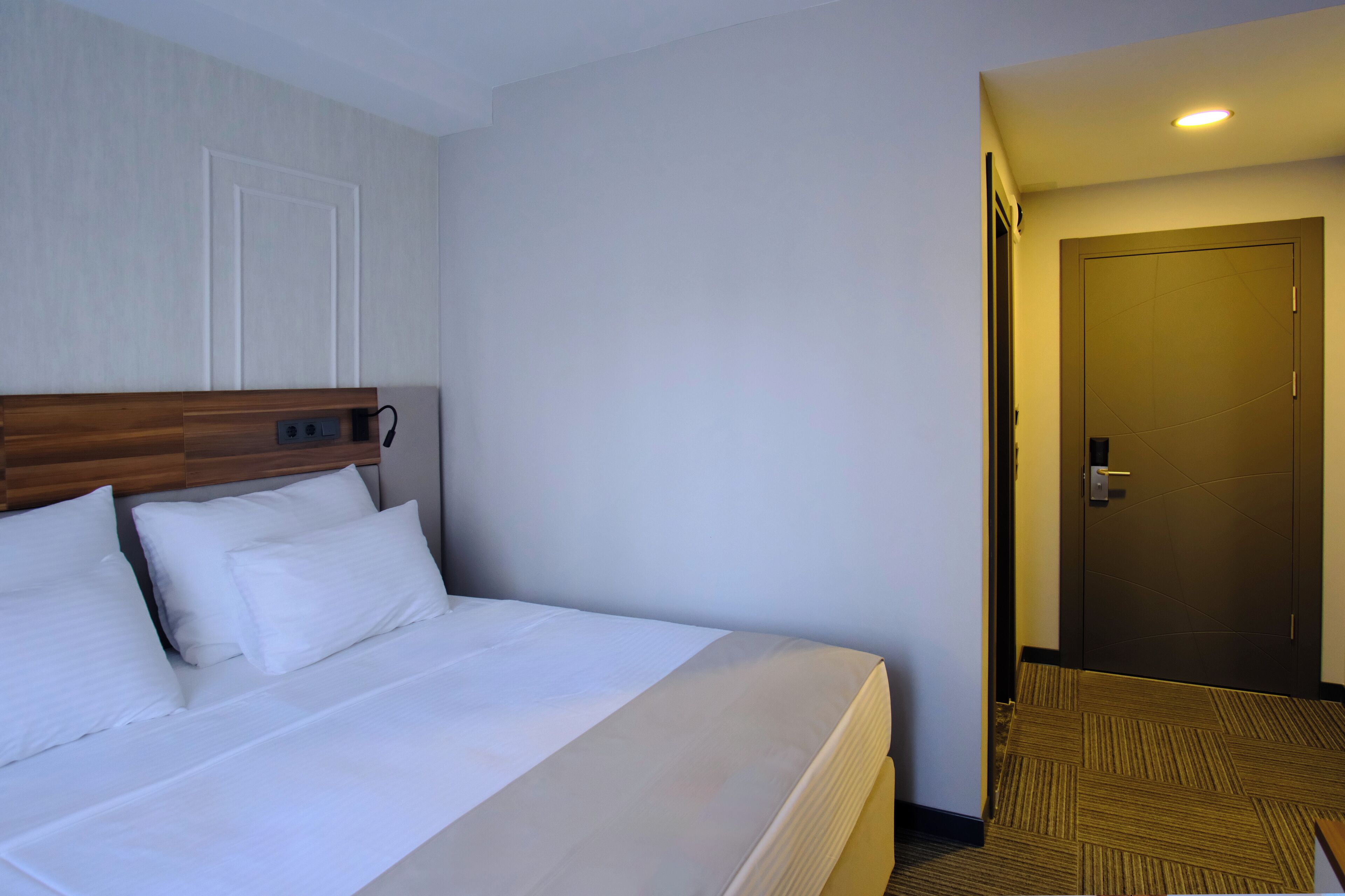 Una habitación de hotel con una cama, cabecero de madera, puerta blanca y suelo alfombrado.