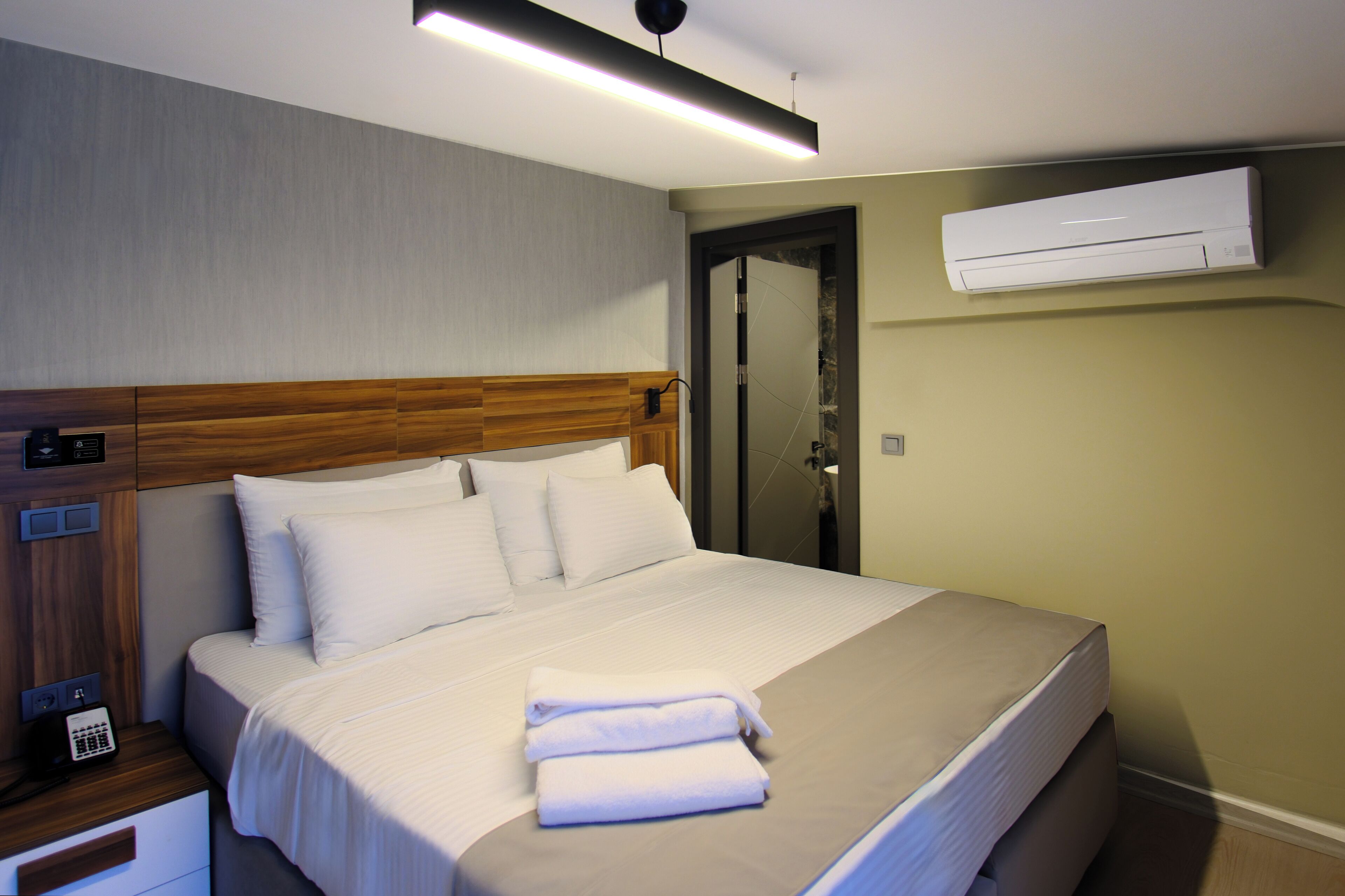 A neatly made bed with white linens and a folded towel, a wooden headboard, a bedside phone, and an air conditioning unit mounted on the wall.
