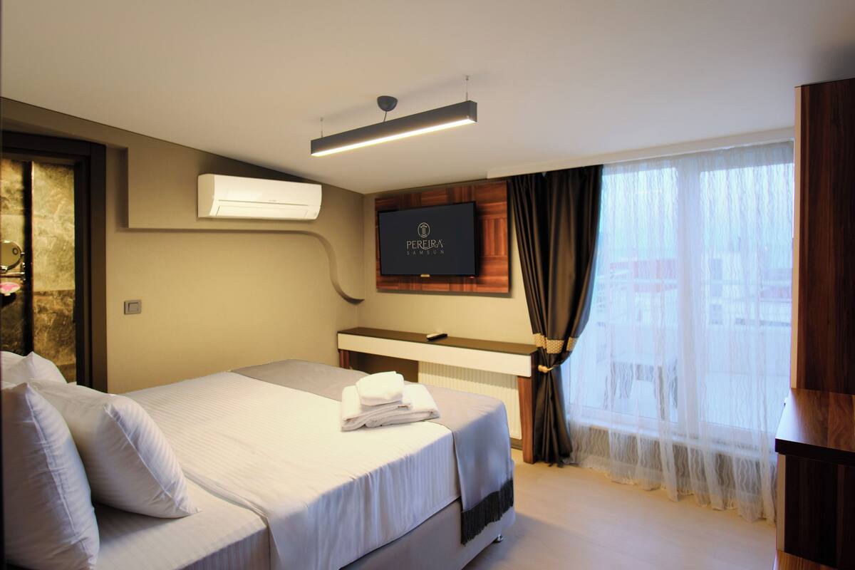A hotel room with a bed, a flat-screen TV mounted on the wall, a desk, and a large window with curtains.