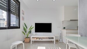 A modern, well-lit room with a flat-screen TV mounted on the wall, a small dining table, a white sofa, and a potted plant.