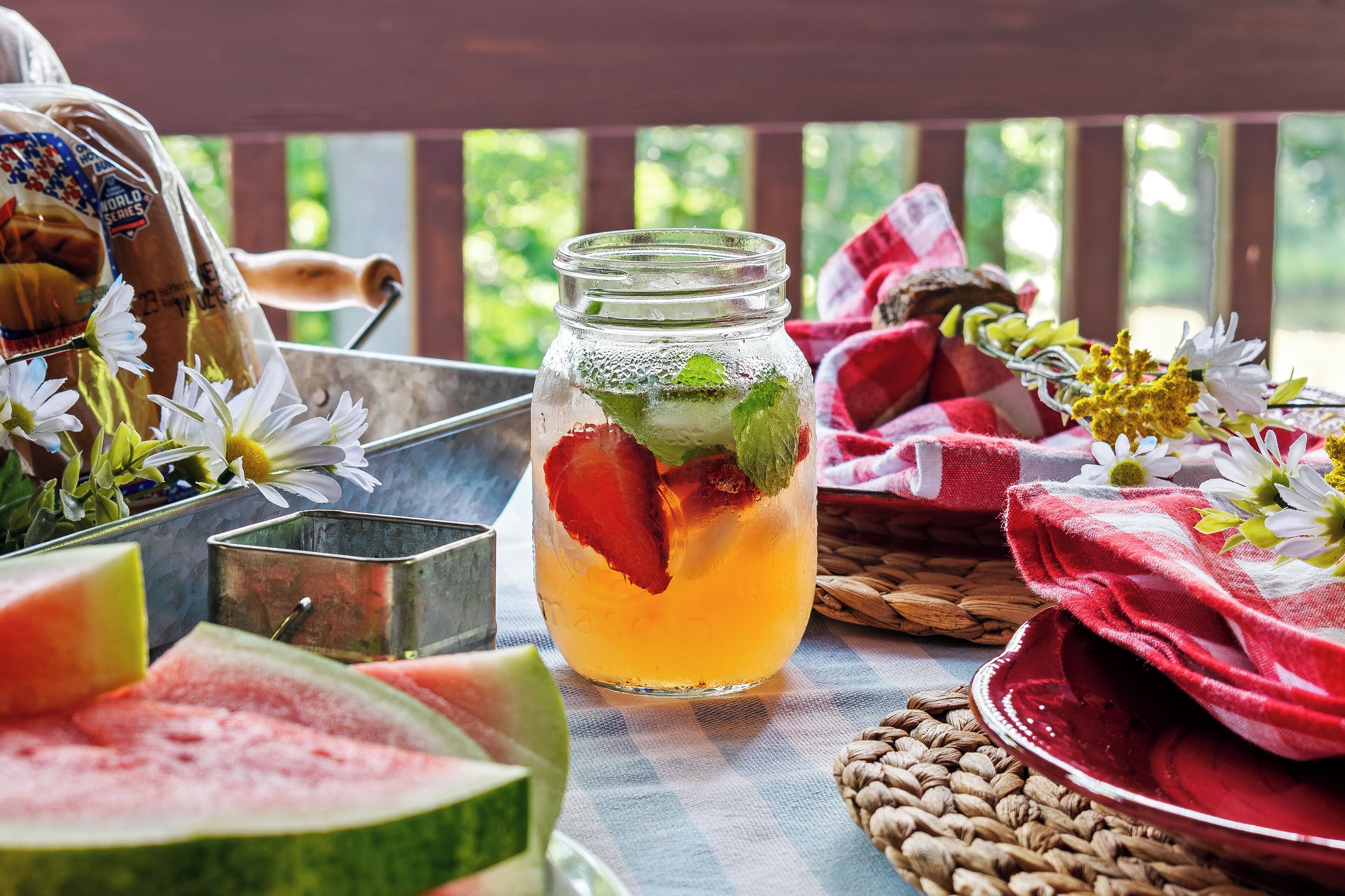 A glass jar with a beverage, surrounded by fresh flowers, a metal container, and a red cloth on a checkered tablecloth.