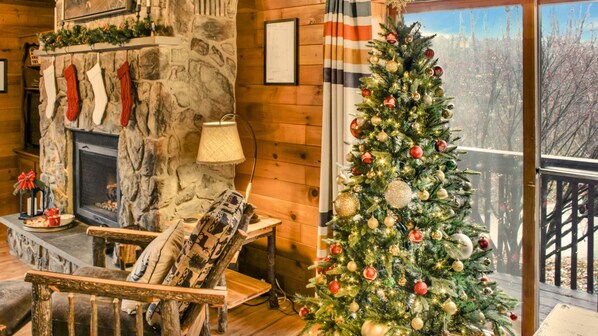 Interior acogedor de una cabaña con chimenea de piedra, un árbol de Navidad decorado y vistas a un paisaje nevado a través de una ventana.
