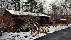 A log cabin with a porch and a smaller attached structure, surrounded by snow-covered ground and trees.