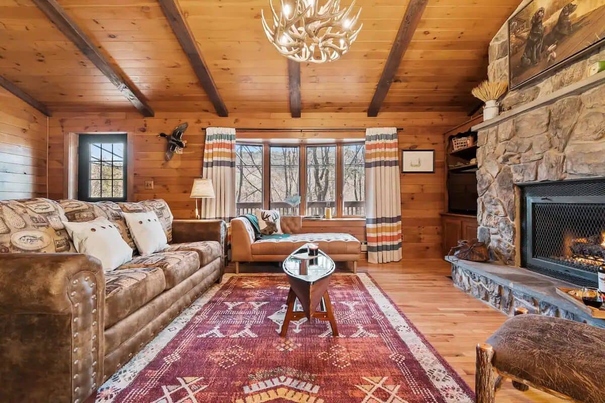 L’intérieur chaleureux d’un chalet avec un plafond en bois, une cheminée en pierre, un canapé agrémenté de coussins à motifs et un tapis.